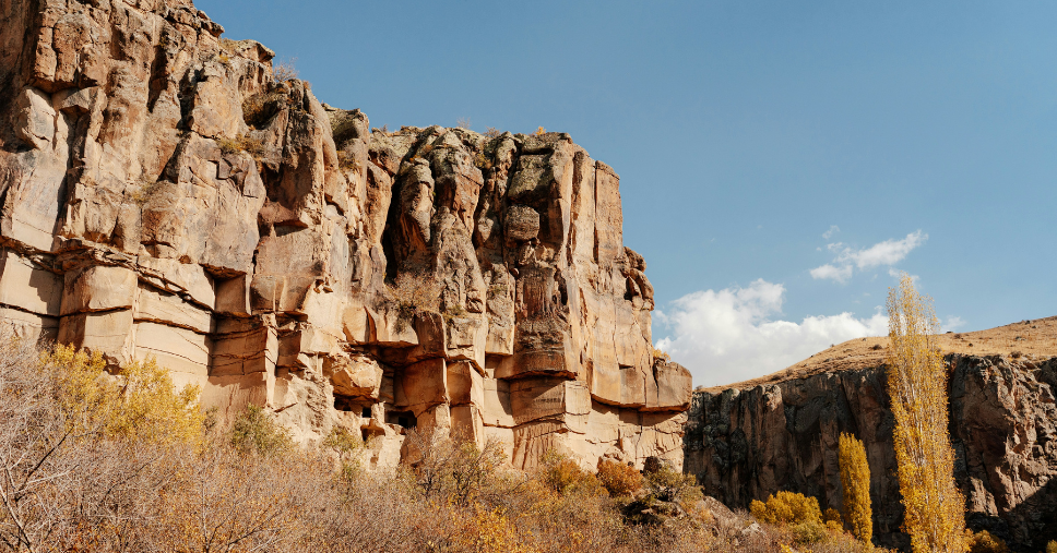 Towering rock cliffs in Ihlara Valley, Cappadocia, surrounded by autumn-coloured trees under a clear blue sky.