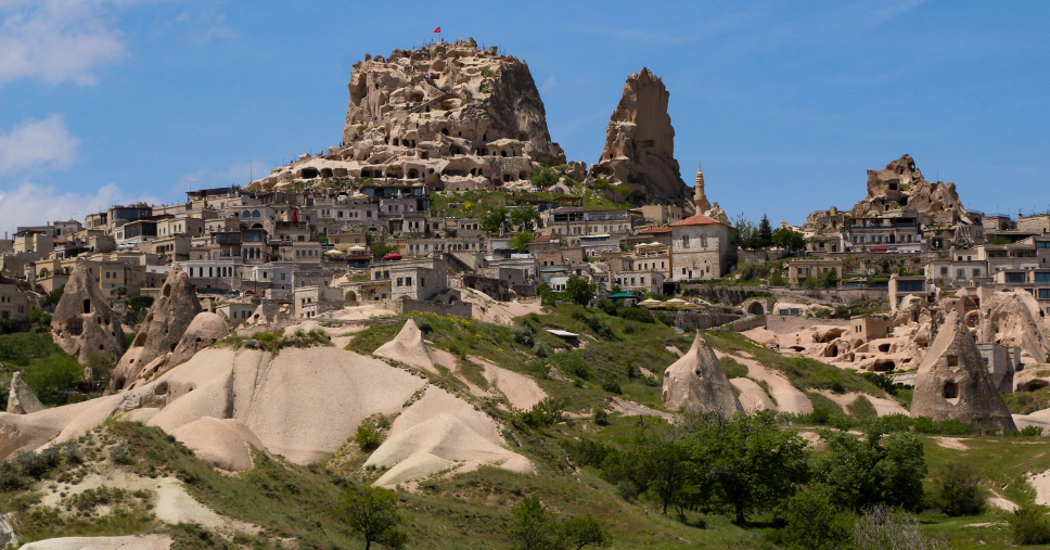 Uchisar Castle perched atop a rocky hill, overlooking a village with stone houses and narrow streets, with the Turkish flag at the summit.